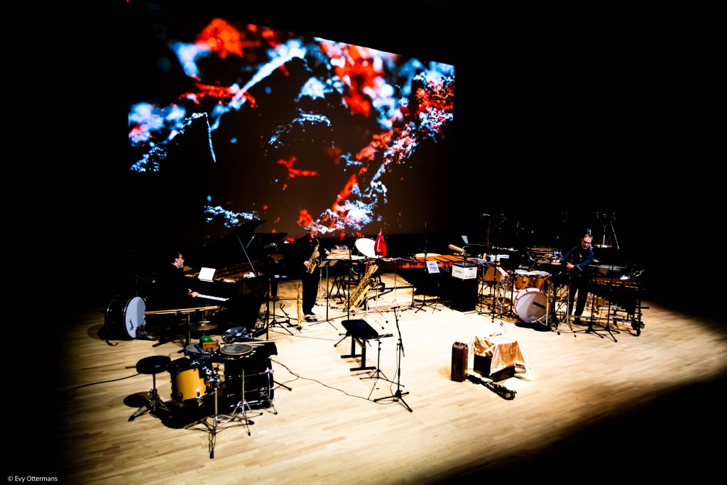 Een muziekensemble dat optreedt op een podium, met een grote projectie aan de achtergrond, die abstracte beelden toont. De muzikanten bespelen verschillende instrumenten, waaronder een piano, saxofoon en percussie.