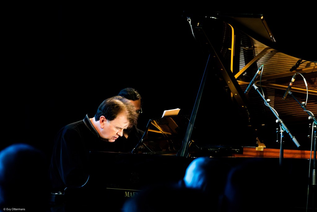 Een pianist in geconcentreerde houding tijdens een uitvoering aan een vleugelpiano, met een donkere achtergrond en enkele microfoons zichtbaar.