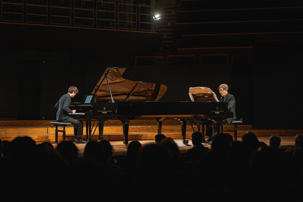 Twee pianisten spelen samen op een podium tijdens een concert, met publiek op de voorgrond en een open piano zichtbaar.