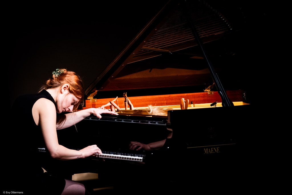 Een pianiste met rode haren speelt intensief op een vleugel, met een focus op de toetsen. Het podium is sfeervol verlicht en de achtergrond is donker.