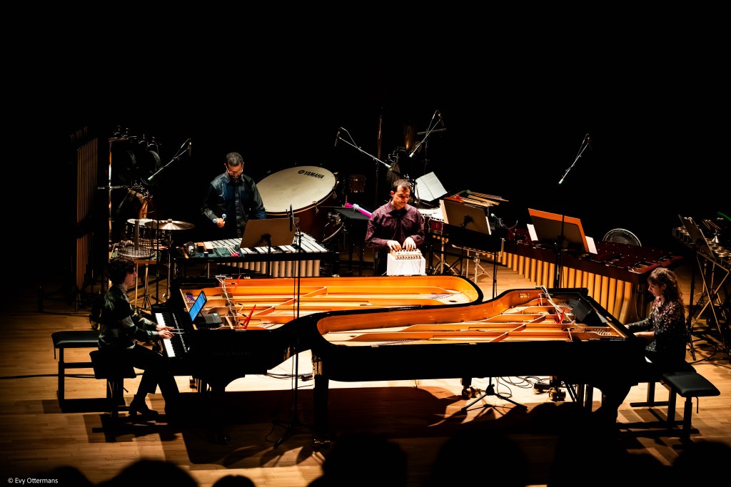 Twee pianisten en twee percussionisten tijdens een live optreden, met diverse muziekinstrumenten op het podium en een focus op moderne klassieke muziek.