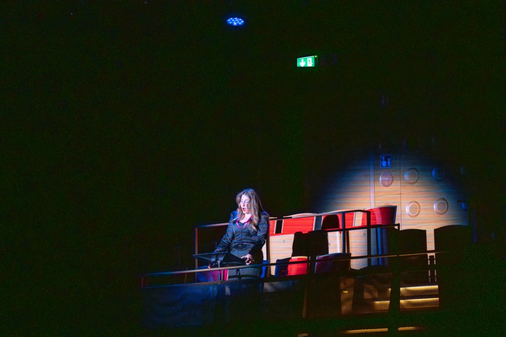 A woman standing on a stage in a dimly lit theatre, wearing a black outfit and speaking, with red chairs in the background.