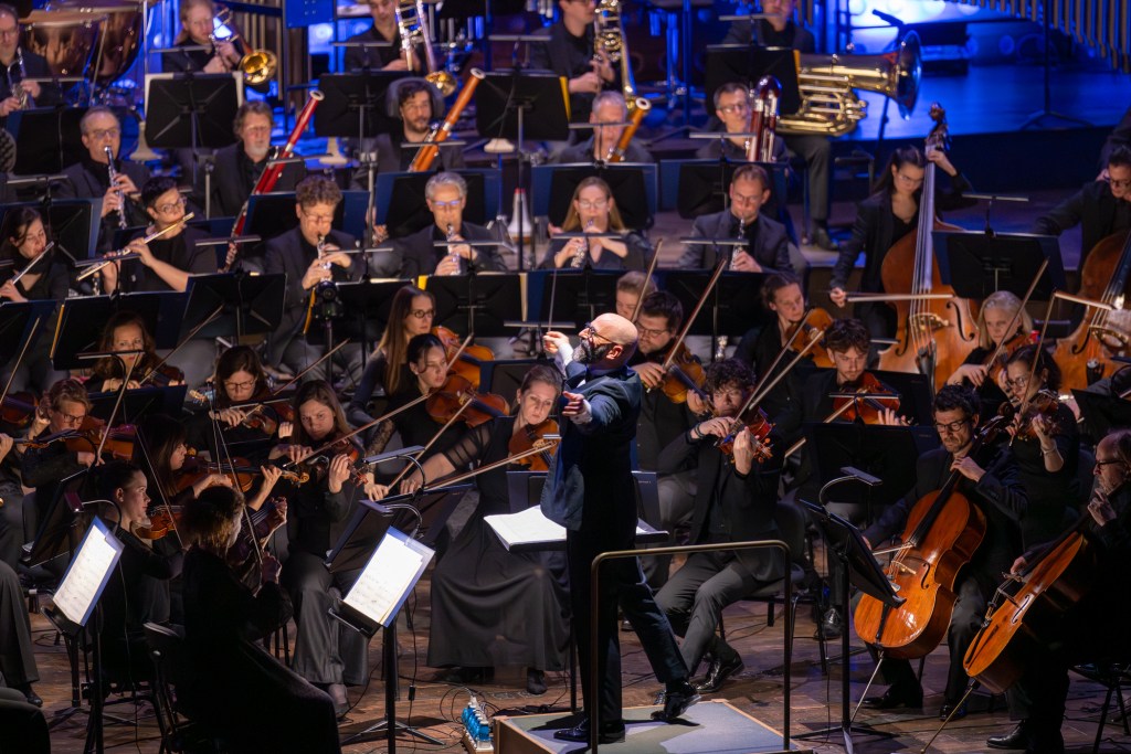 A conductor leading a large orchestra performing on stage, with musicians playing various instruments like violins, flutes, and brass, during a live concert.